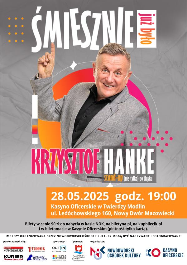 Mężczyzna w czarnej koszuli i szarej marynarce z uniesioną prawą ręką z palcem wskazującym do góry.