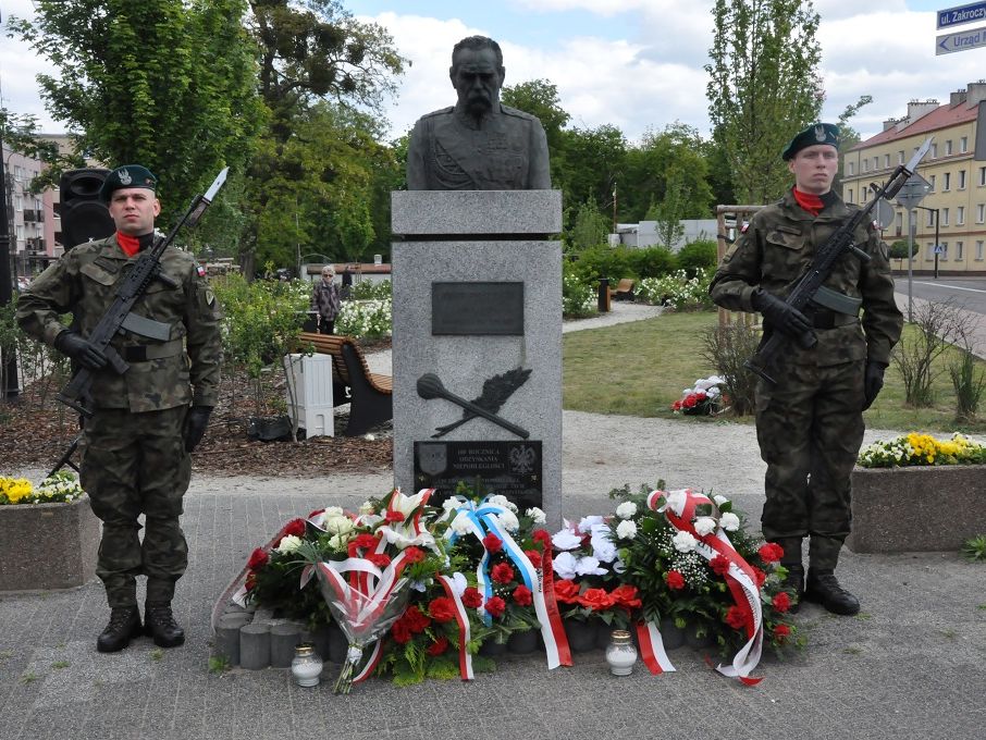 Dwóch żołnierzy z karabinami na piersiach stojących po obydwu stronach pomnika, poniżej wiązanki kwiatów.