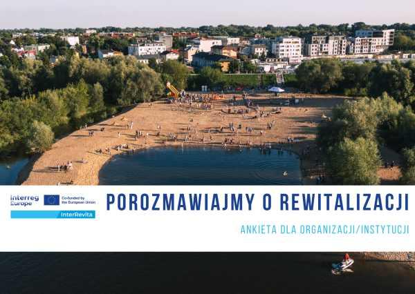 Na piaszczyste plaży przy wodzie kilkadziesiąt osób. W oddali wał przeciwpowodziowy i budynki mieszkalne.