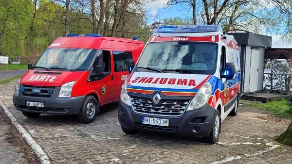 Czerwony samochód z białym napisem STRAŻ oraz niebieskimi lampami sygnalizacyjnymi. Obok biały samochód z czerwonym napisem AMBULANS, niebiesko-czerwonym pasem z przodu i boku samochodu.