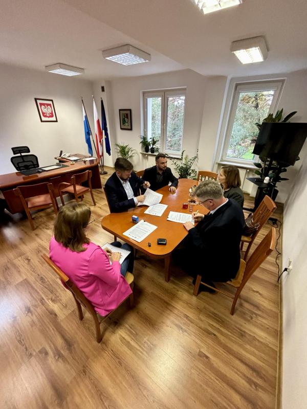 Przy stole siedzi trzech mężczyzn i dwie kobiety. Dwóch mężczyzn podpisuje dokumenty.