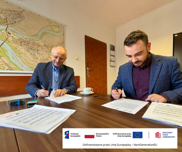 Dwóch mężczyzn w niebieskich marynarkach podpisuje dokumenty na brązowym stole.