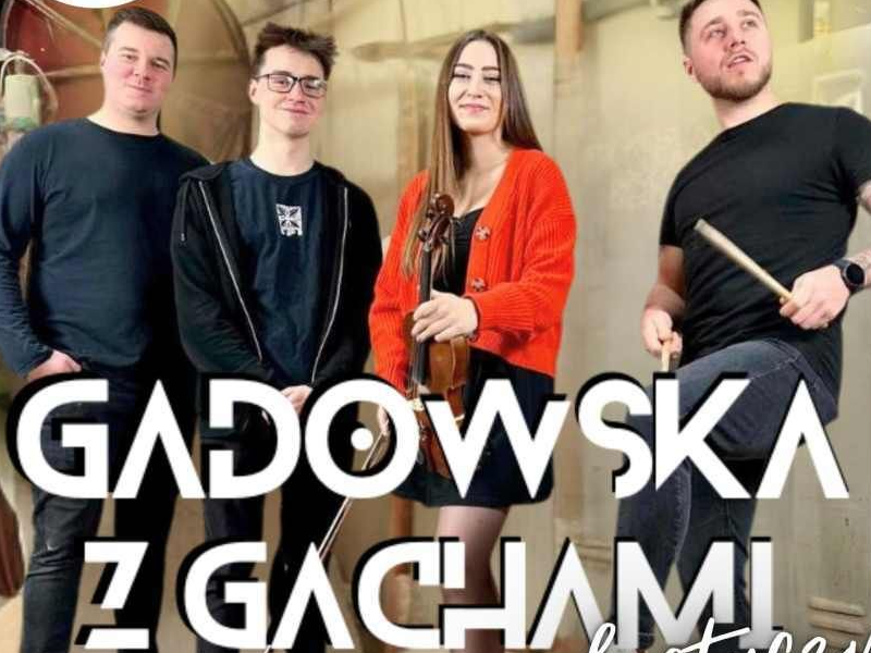 Kobieta trzyma w dłoniach skrzypce i smyczek. Obok niej stoi trzech mężczyzn, jeden z nich z pałeczkami perkusyjnymi.