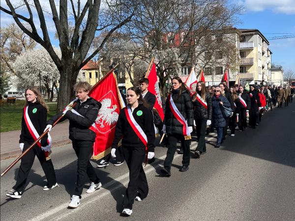 Młodzież przepasana biało-czerwonymi szarfami idąca ze sztandarami na drewnianych drzewcach.