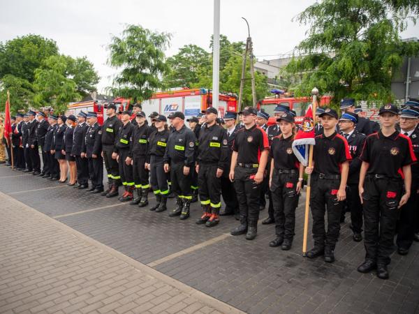Kilka kobiet i kilkudziesięciu mężczyzn w mundurach strażackich stojących na placu na tle wozów strażackich.