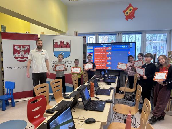 Dzieci stojące z dyplomami w rękach. Po lewej stronie stoi mężczyzna. Za nimi dwa rollupy i duży monitor.