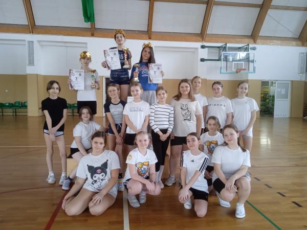 Dziewczynki w strojach sportowych na sali gimnastycznej, trzy z nich w koronach, trzymające w rękach puchary i dyplomy.