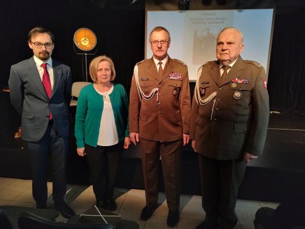 Po lewej stronie kobiety stojący mężczyzna w garniturze, po jej prawej stronie dwóch mężczyzn w mundurach wojskowych.