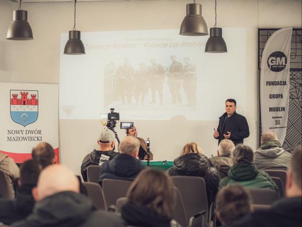 Mężczyzna mówiący do grupy osób, za nim wyświetlony slajd na ścianie, obok roll-up.