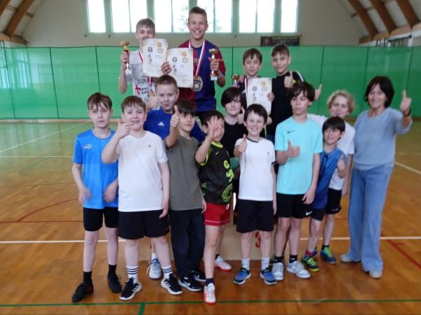 Grupa chłopców stojących na parkiecie i podium, trzech z medalami, pucharami i dyplomami, obok kobieta.