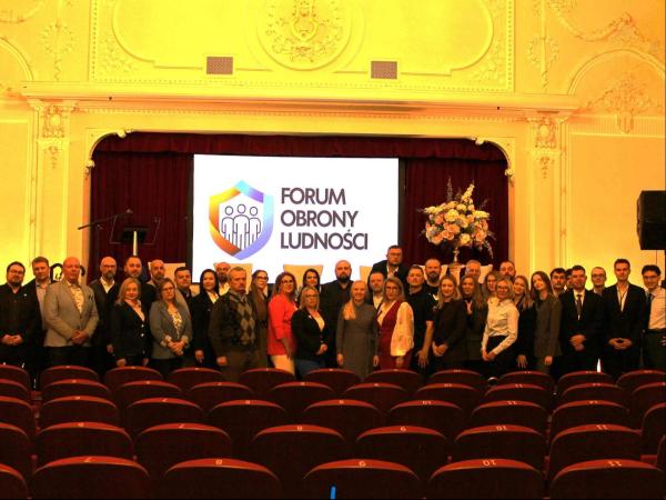 Uczestnicy Forum stojący obok siebie, za nimi slajd Forum Obrony Ludności.