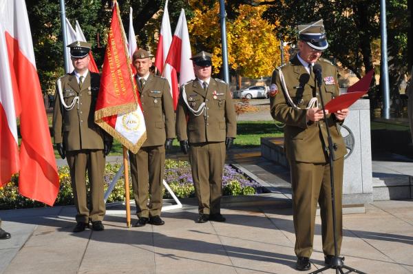 Trzech żołnierzy stojących obok siebie. środkowy trzyma sztandar. Z boku oficer przy mikrofonie na statywie.